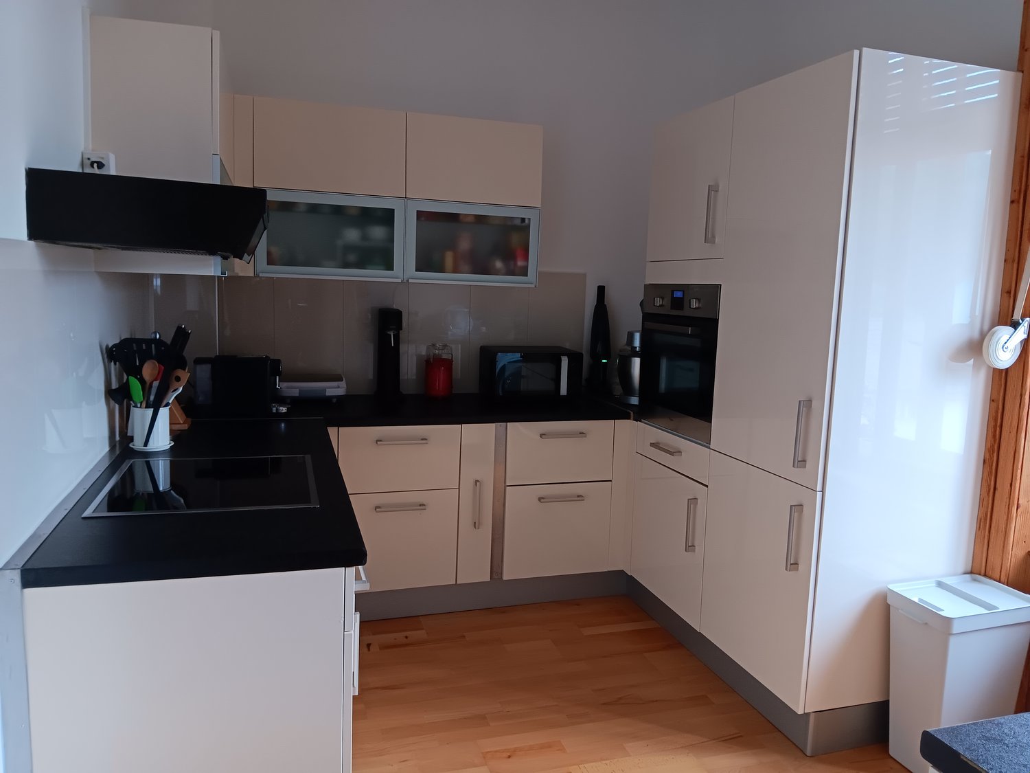 Modern kitchen with beige cabinets, black countertop, built-in appliances including a dishwasher, and hardwood flooring.