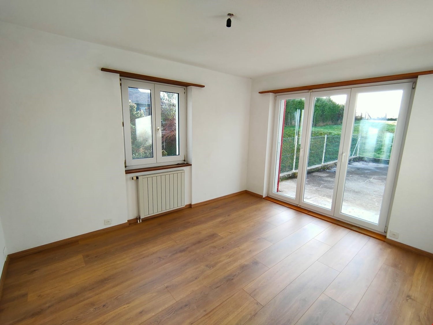 Spacious living room with large windows overlooking the outdoor area, hardwood flooring, and a radiator on the wall.