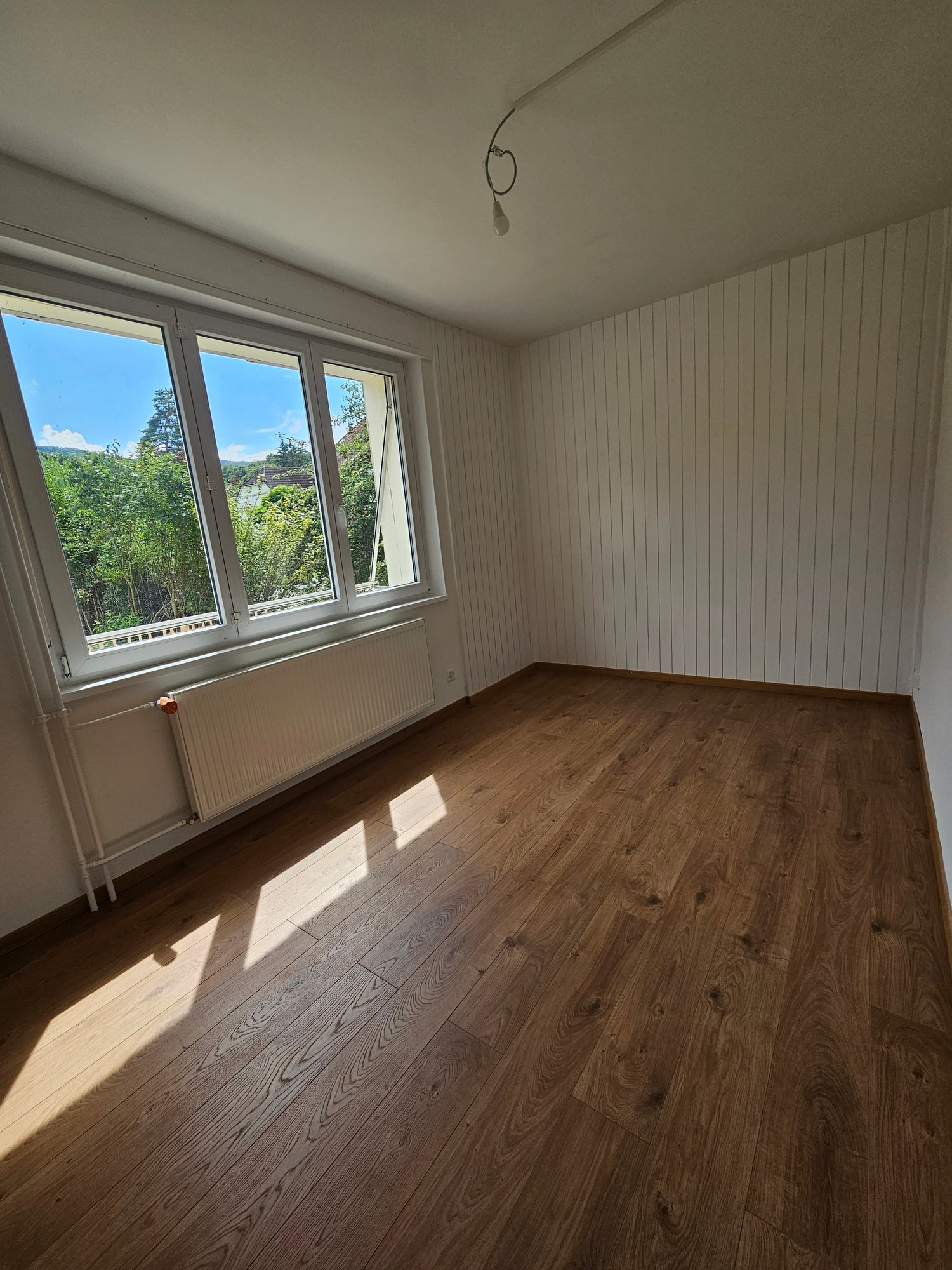 This image shows an empty room with hardwood flooring and white paneled walls. The room has large windows that provide a view of the surrounding greenery outside. The room appears to be a bedroom or living space with a simple and minimalist design.