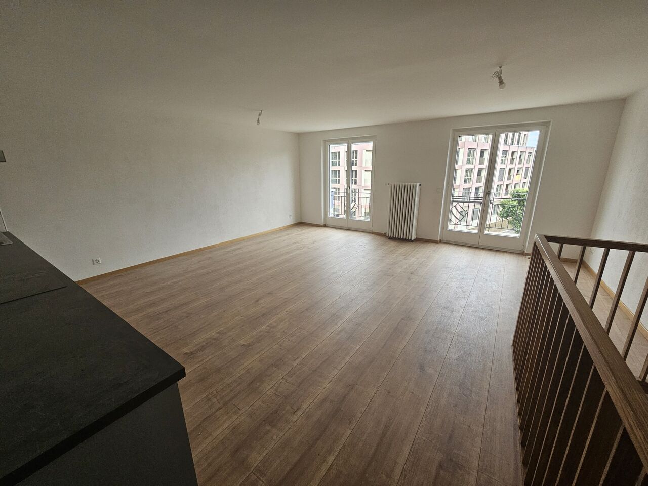 Spacious living room with hardwood flooring, large windows, and a radiator. The room has an open layout with a balcony or terrace access.