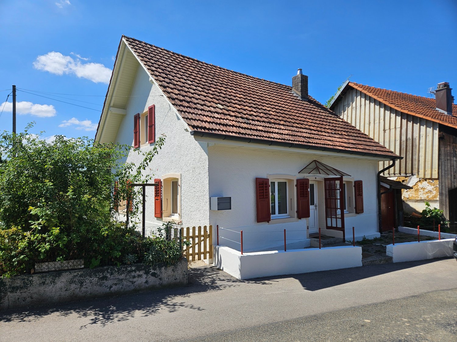 Two-story house with a white exterior, red tile roof, and red shutters. The house has a porch with a wooden fence and steps leading up to the entrance. There are trees and greenery surrounding the house.