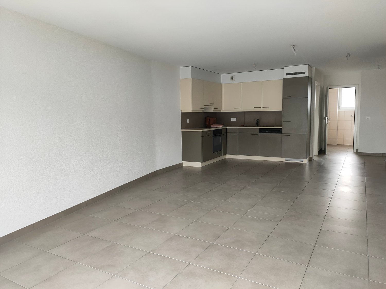 The image shows a spacious open-plan apartment with a modern kitchen and living area. The kitchen features beige cabinets and a black appliance, while the living area has a tiled floor. The apartment appears to have a clean and minimalist design with whit