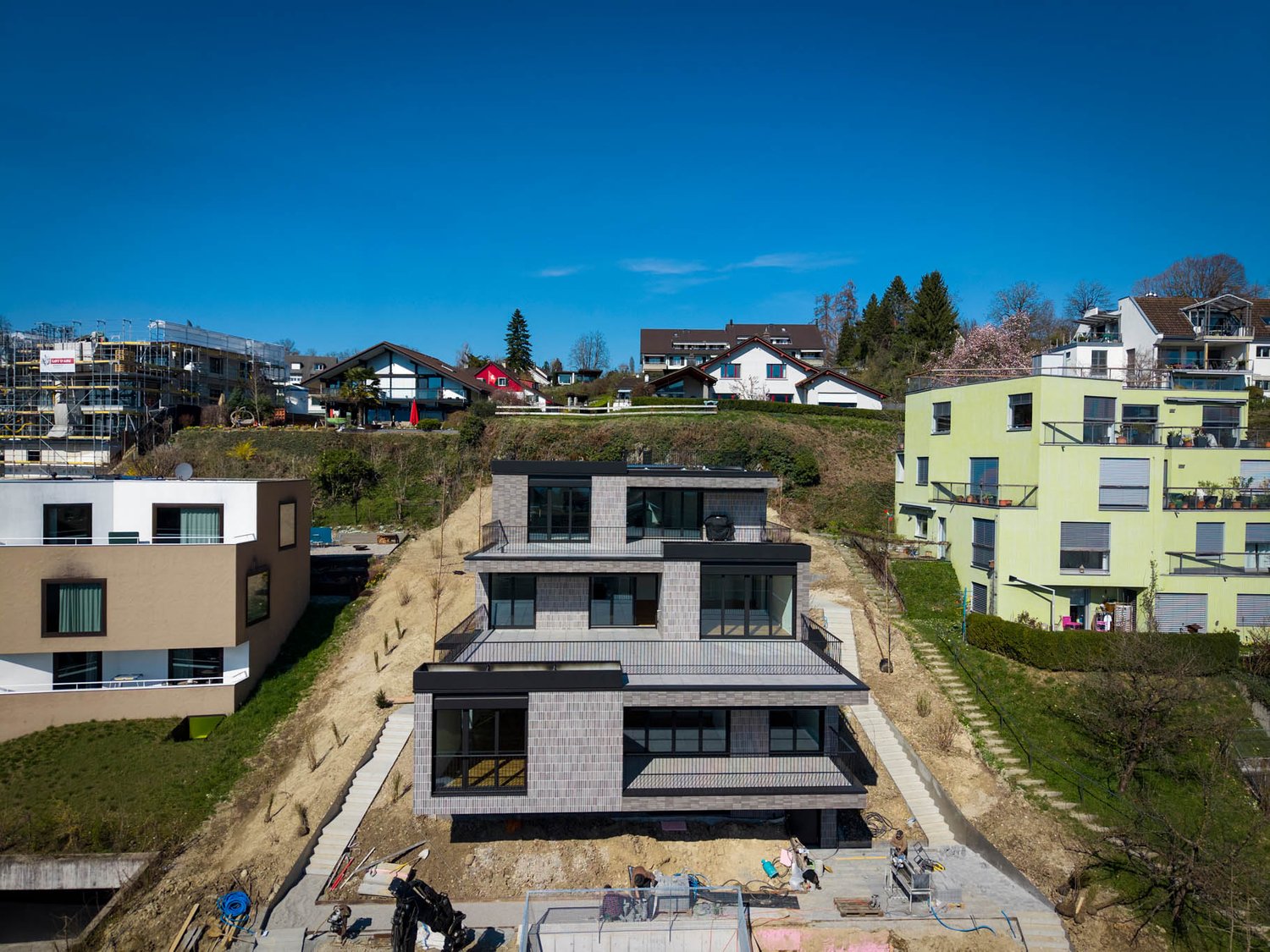 Modern house under construction, multiple levels, large windows, balconies, surrounded by other houses and greenery