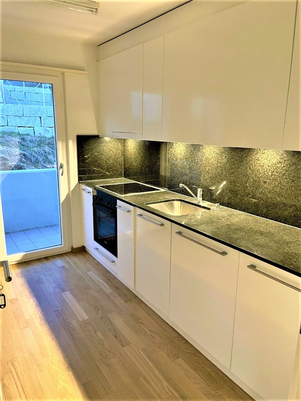 Modern kitchen with white cabinets, wooden floor, granite countertops, integrated sink, built-in oven, sliding glass door to the balcony.