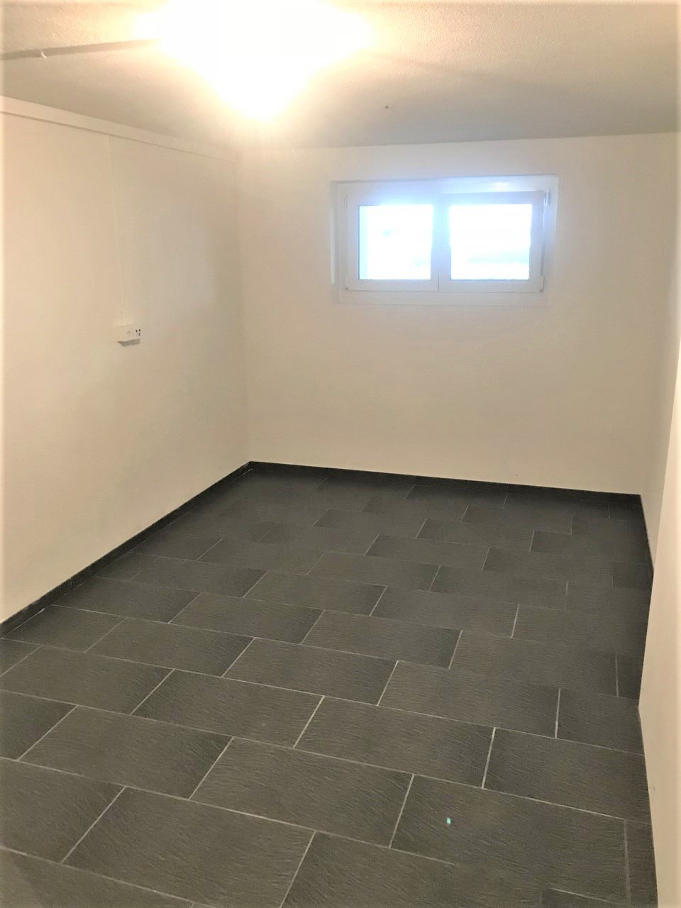 Empty room with white walls, grey tiled floor, one window.