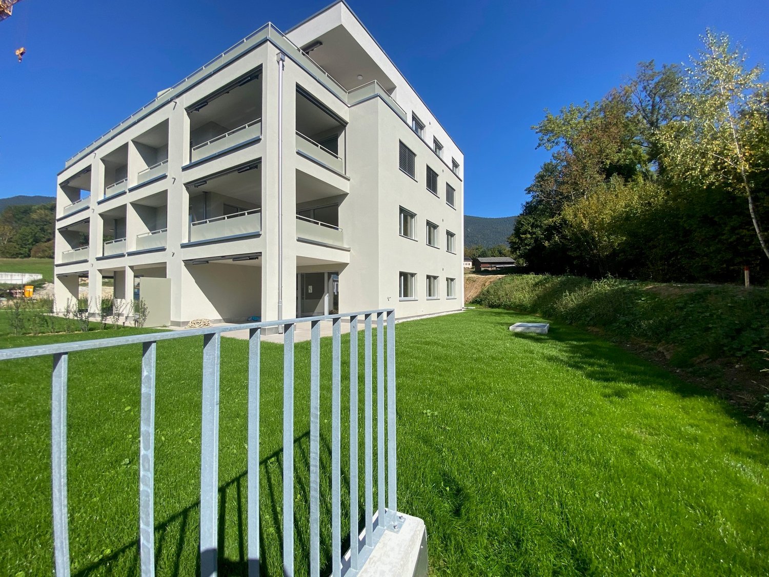Modern white apartment building with multiple balconies, on multiple floors, set in a well-maintained lawn with mature trees and a scenic mountain range in the background