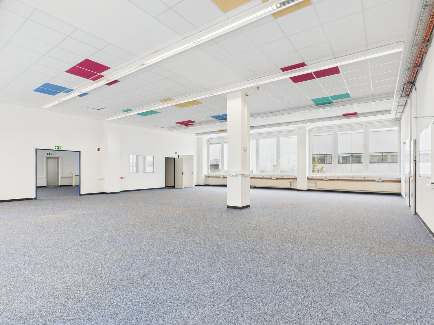 Empty room with gray carpeted floor, white walls, two sets of double doors, large windows, central pillar