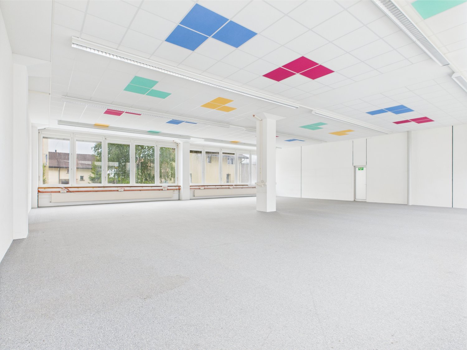 large empty room, white walls, large windows, grey floor, square colored ceiling panels