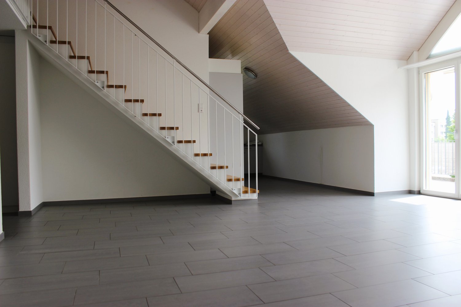 Spacious open-plan living area with a staircase leading to an upper level. The room has white walls, a vaulted ceiling with wooden beams, and a large window providing natural light. The floor is covered with gray tile or laminate flooring.