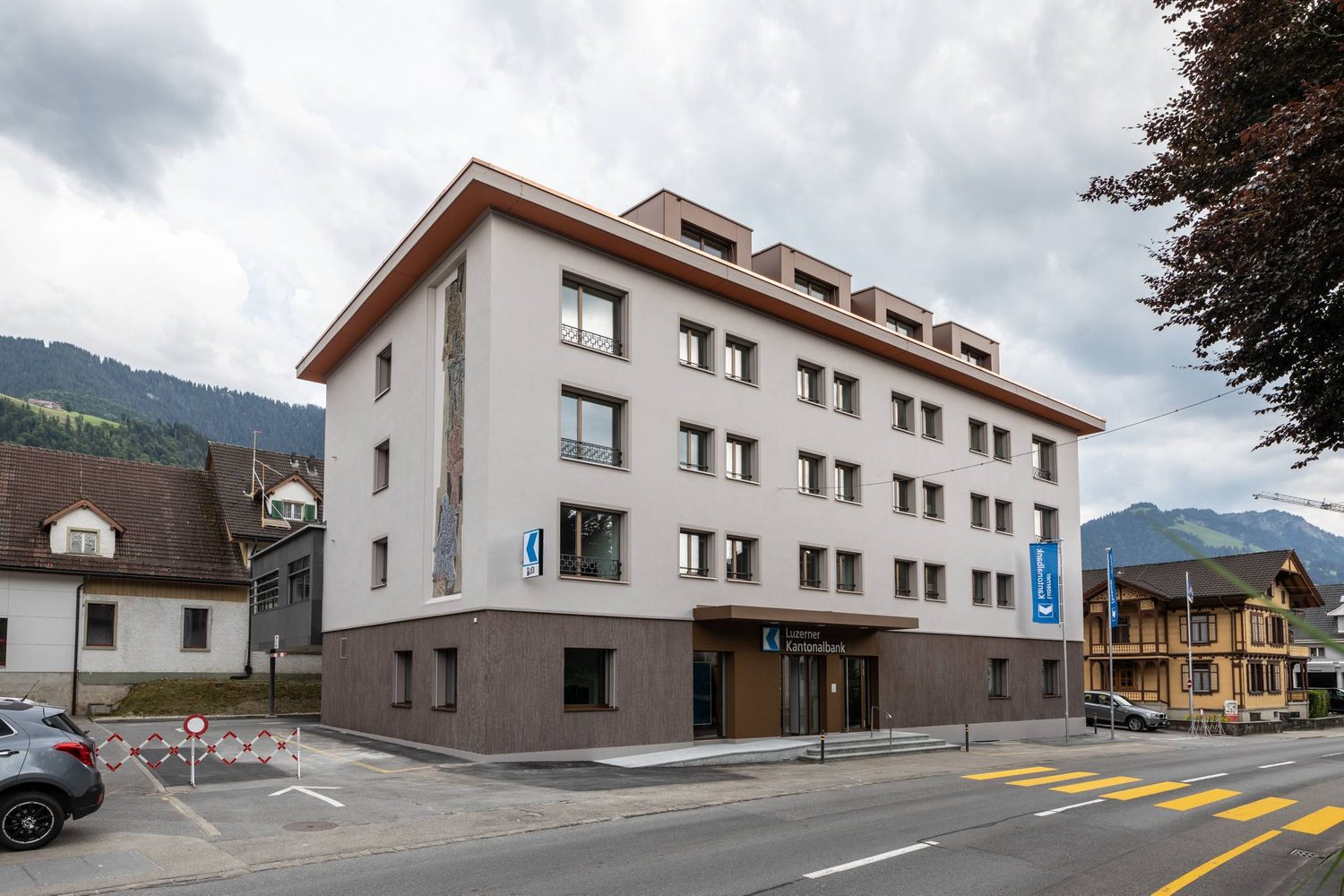 Large white building with brown base, multiple windows, balcony with railings, multiple floors, located in a town with mountains in the background.