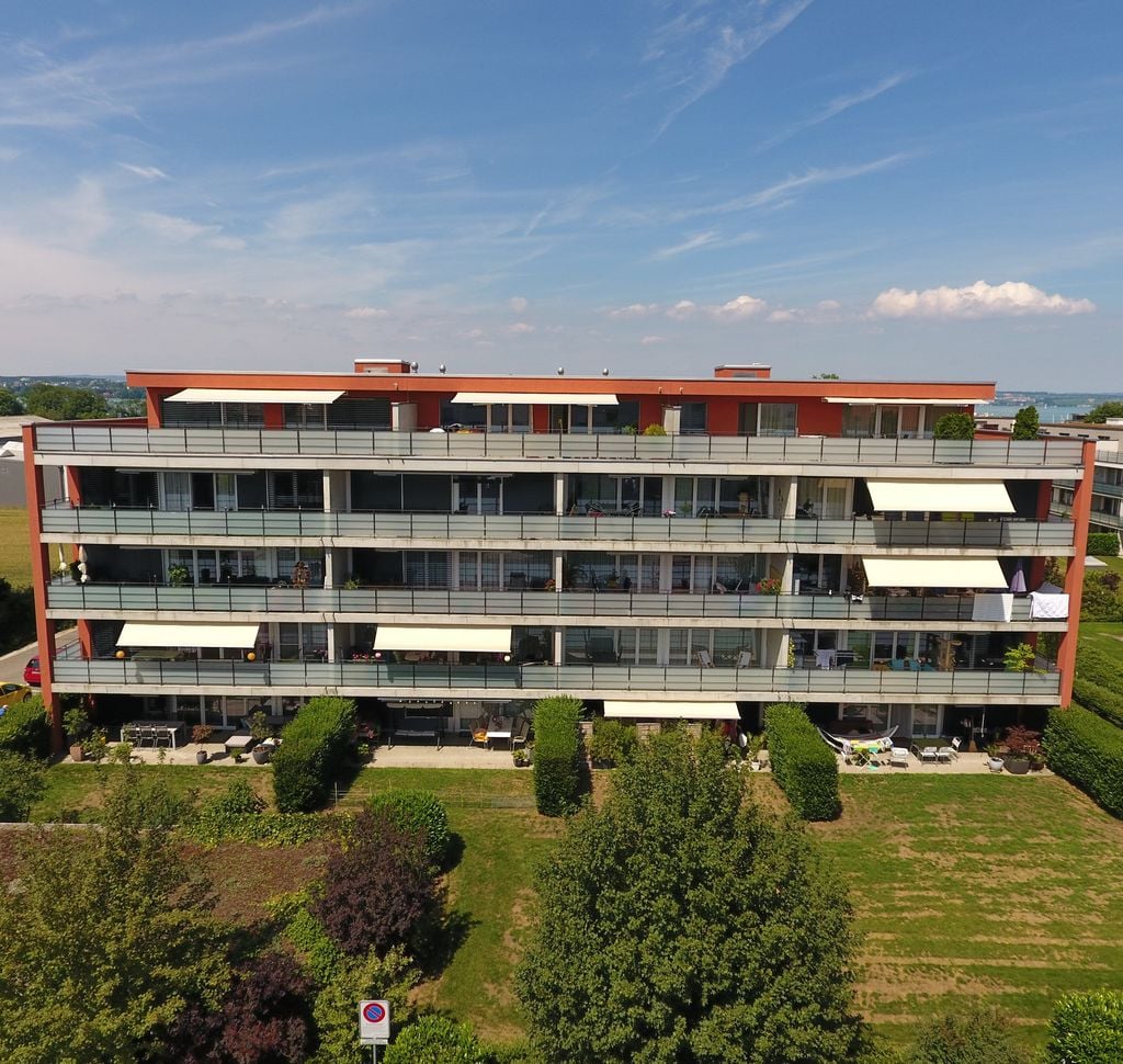Red apartment building with multiple floors, many balconies with awnings, gardens, grass, and trees around