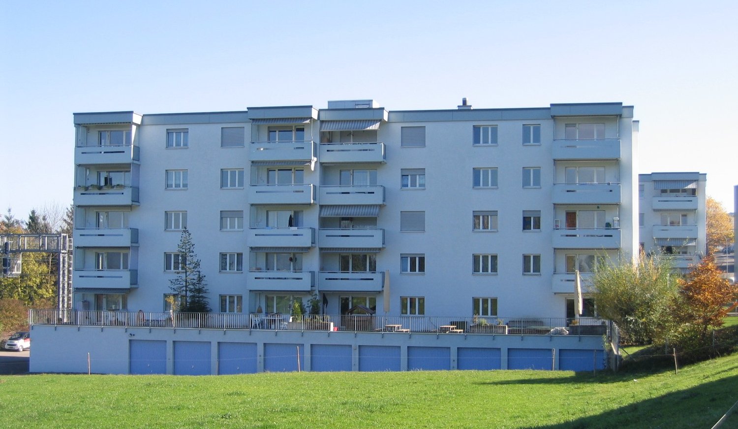 6-story apartment building with white walls and blue garage doors, multiple balconies, ground floor with entrance and small garden, parking space