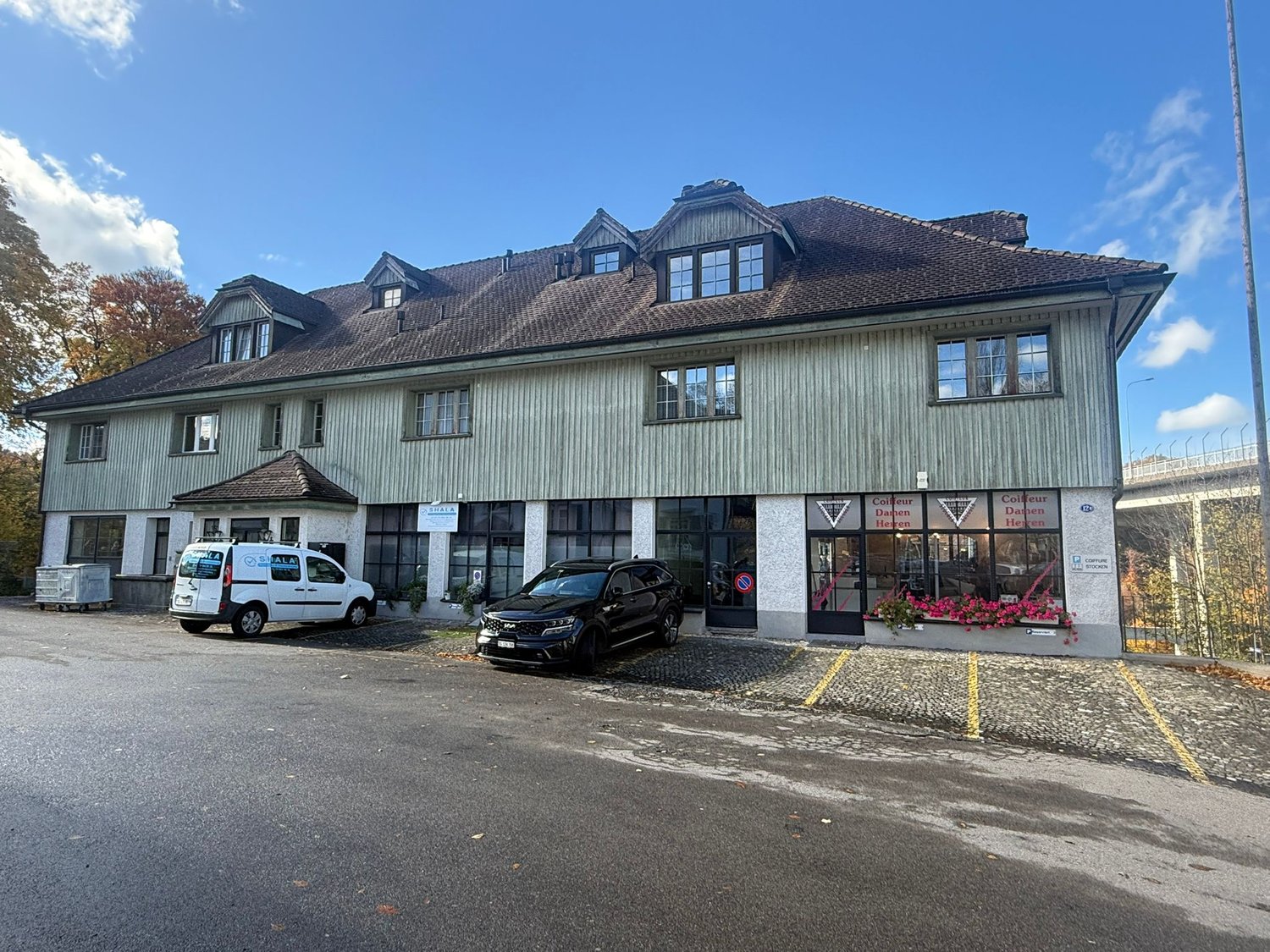 three story building, brown roof, double windows, two cars parked, front garden with plants and flowers