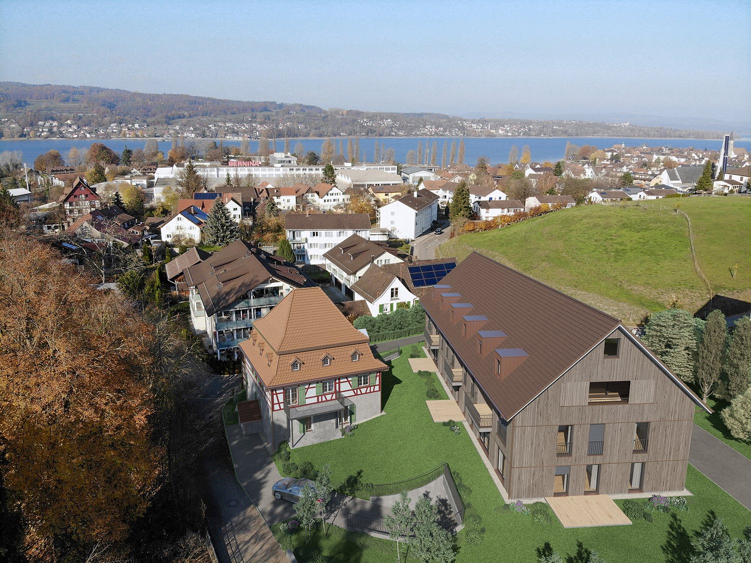 Aerial view of a group of residential houses with brown roofs, a car parked, greenery, and a lake in the distance.