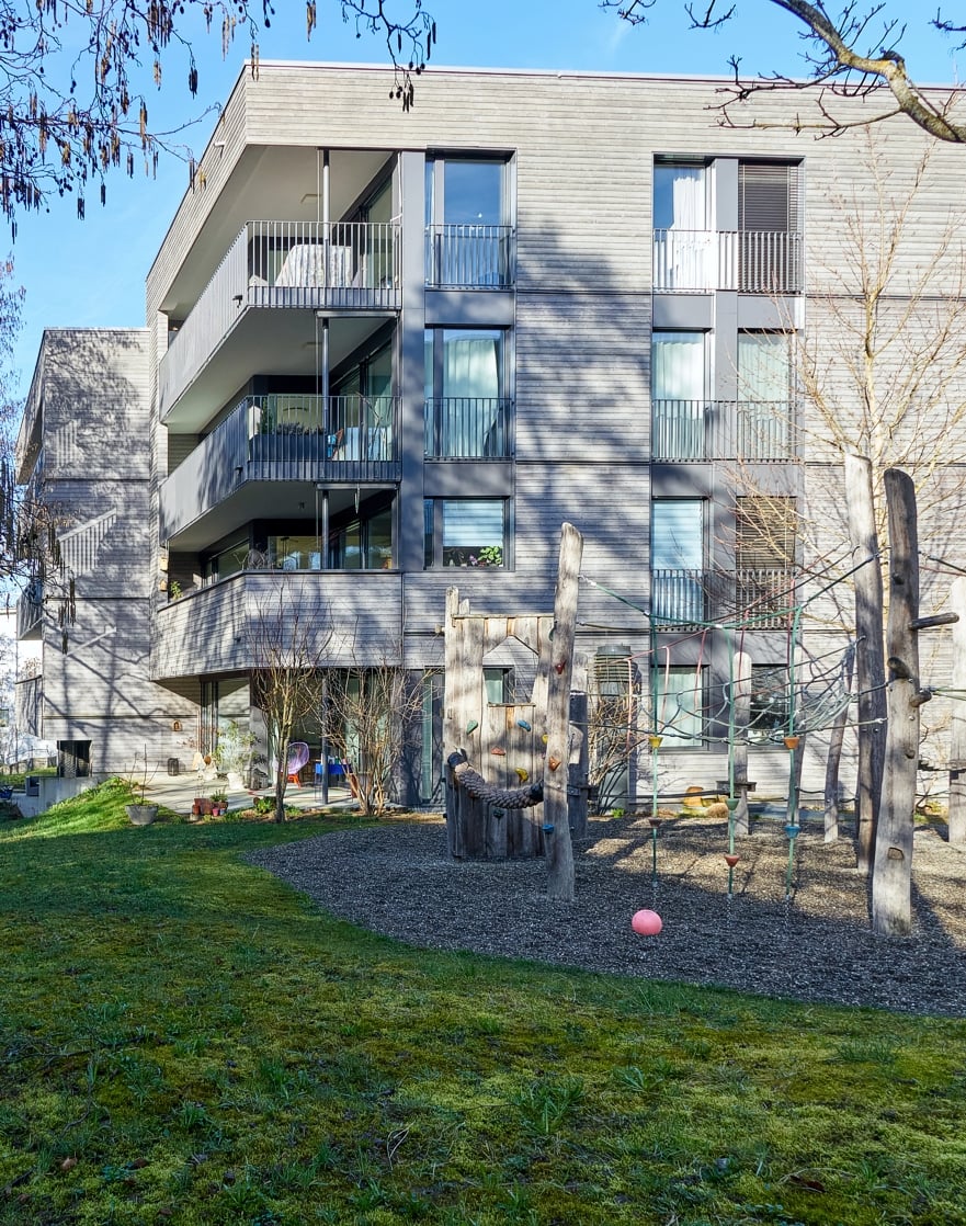 Modern apartment building, concrete exterior, glass windows, balconies on multiple floors, greenery around