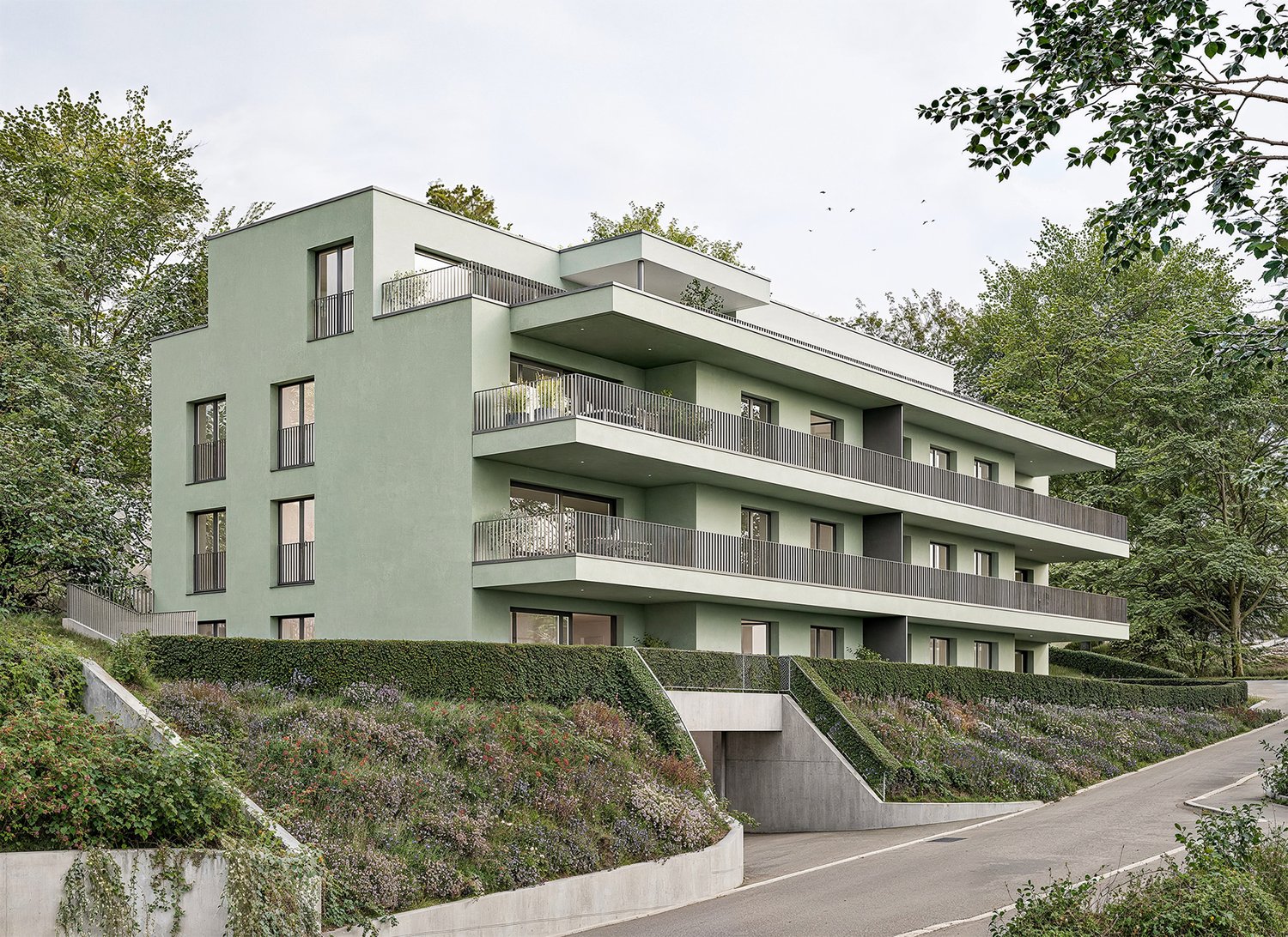 4-story modern apartment building with balconies, surrounded by trees and greenery, with a sloped driveway leading up to the building.