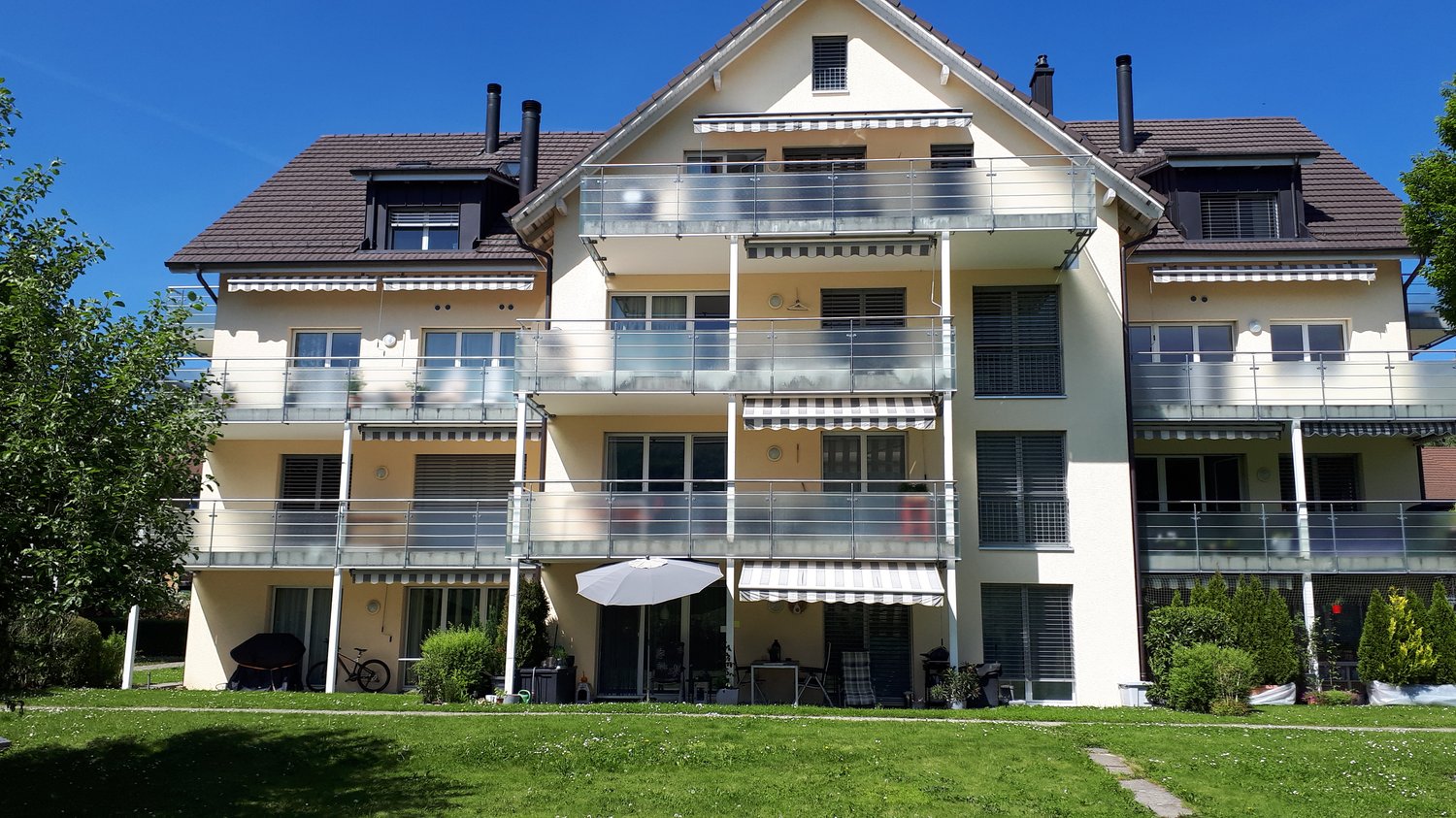 two-story building, balconies, glass railings, awnings, green lawn, plants, bicycle, table with chairs