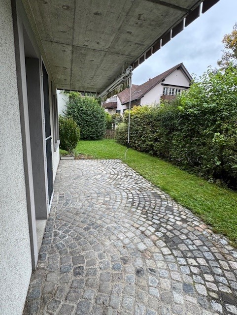 The image shows a raised ground floor with a paved entrance walkway and covered awning. Green grass and plants on both sides of the walkway. A house with a brown roof can be seen in the distance.