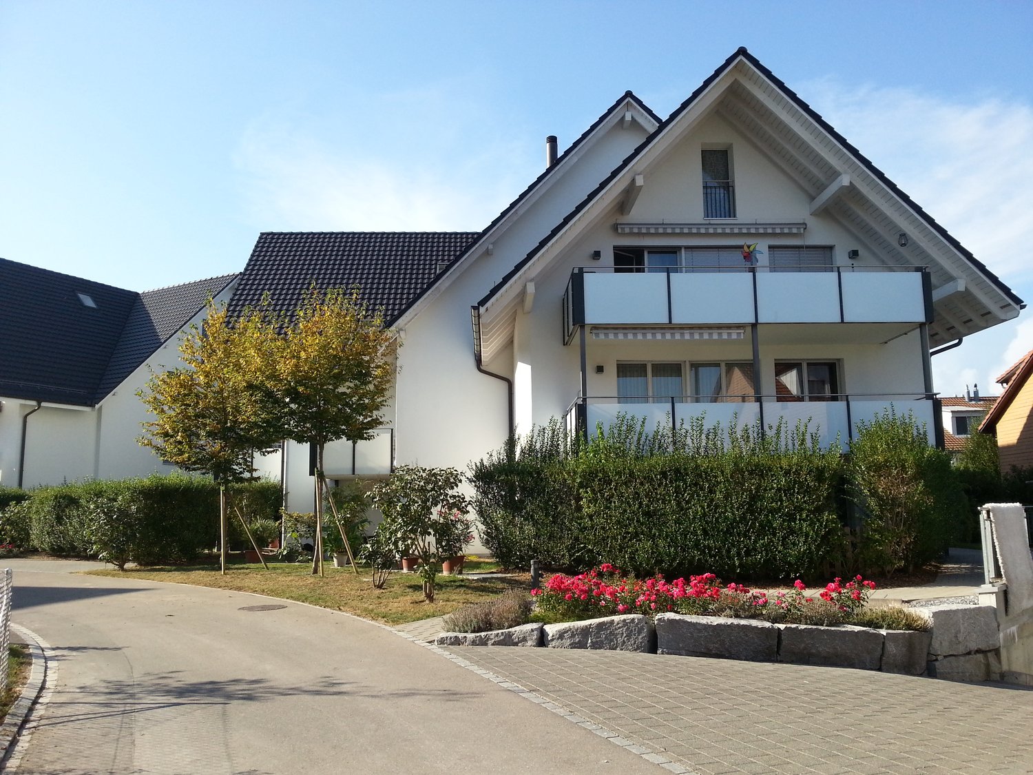 White house, two stories, black roof, balconies, paved driveway, flowers, hedges, bushes