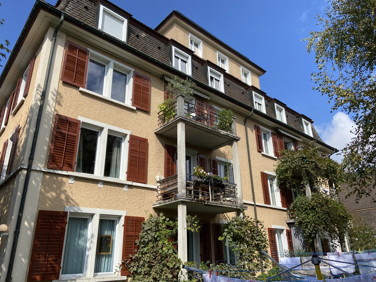 Multi-story building, red shutters, balconies with potted plants and seating, beige exterior, window frames, plants hanging from balconies