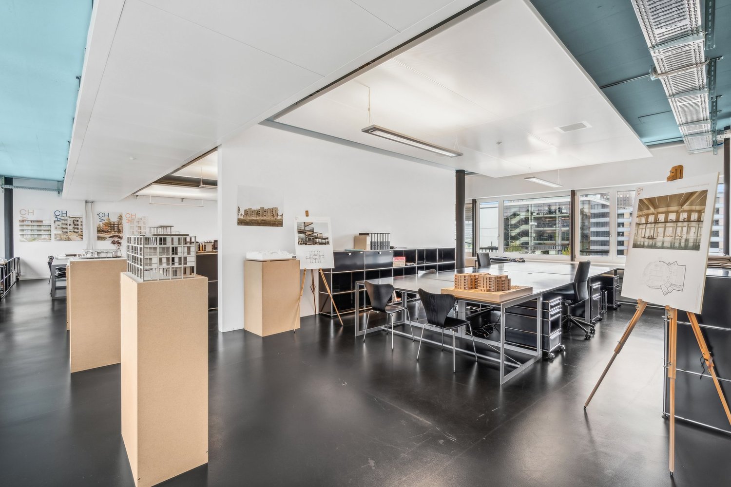 Large open-plan office space with high ceilings, white walls, and concrete flooring. The space contains various workstations, desks, and chairs, as well as architectural models and displays on shelves. There is a view of the city through the large windows
