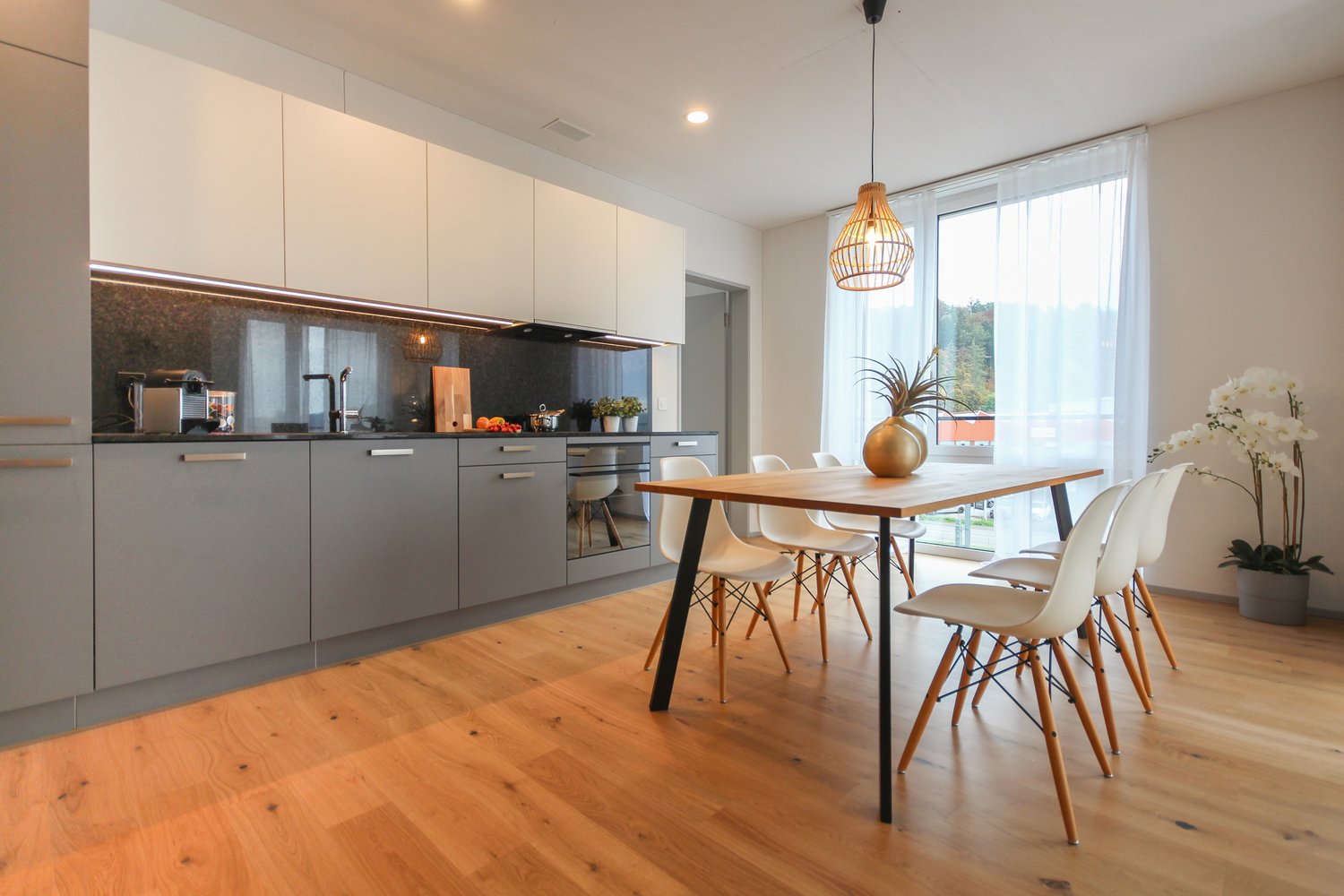 kitchen with wooden table, white chairs, modern cabinets, island, large window with curtains, counter with coffee machine and blender, hanging lamp