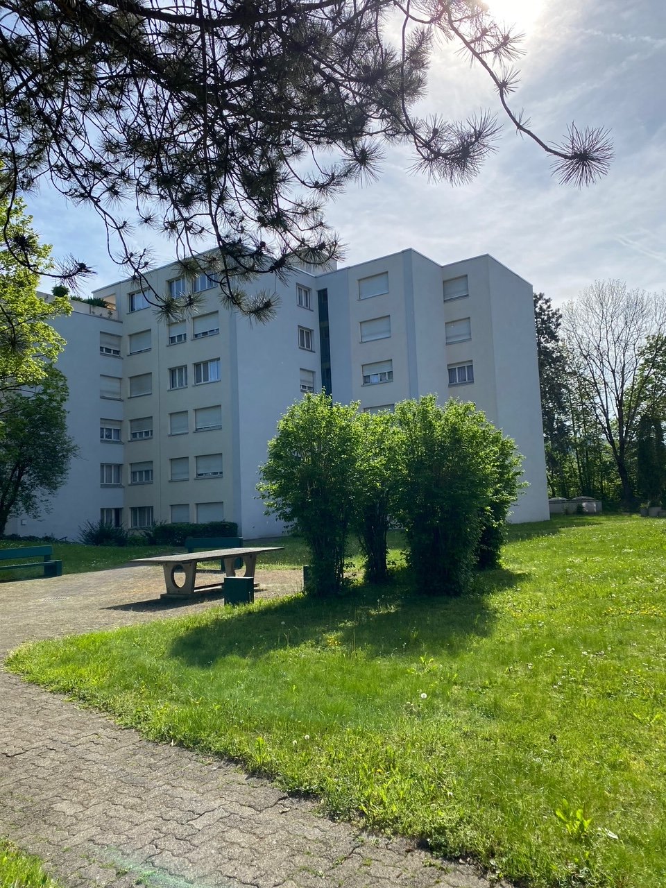 large white apartment building, many windows, balcony, garden, trees, bench