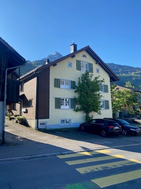 3-story residential building with a traditional Swiss architectural style, featuring a gabled roof, shuttered windows, and a balcony. There is a parking area in front of the building.