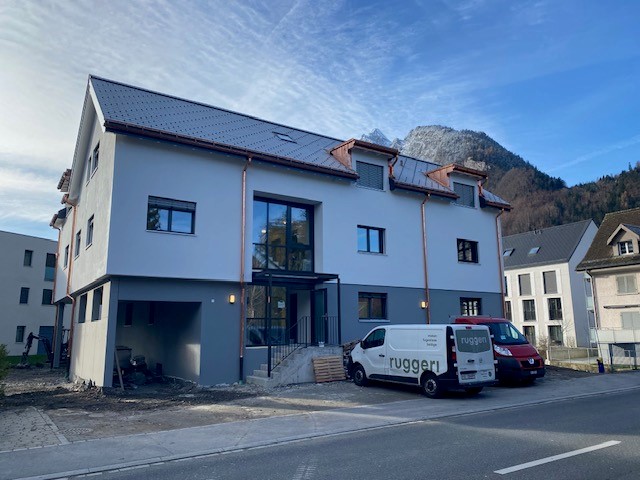 A two-story building with a black roof, two vans parked in front, and construction equipment on the left. It is located in a neighborhood with mountains in the distance.