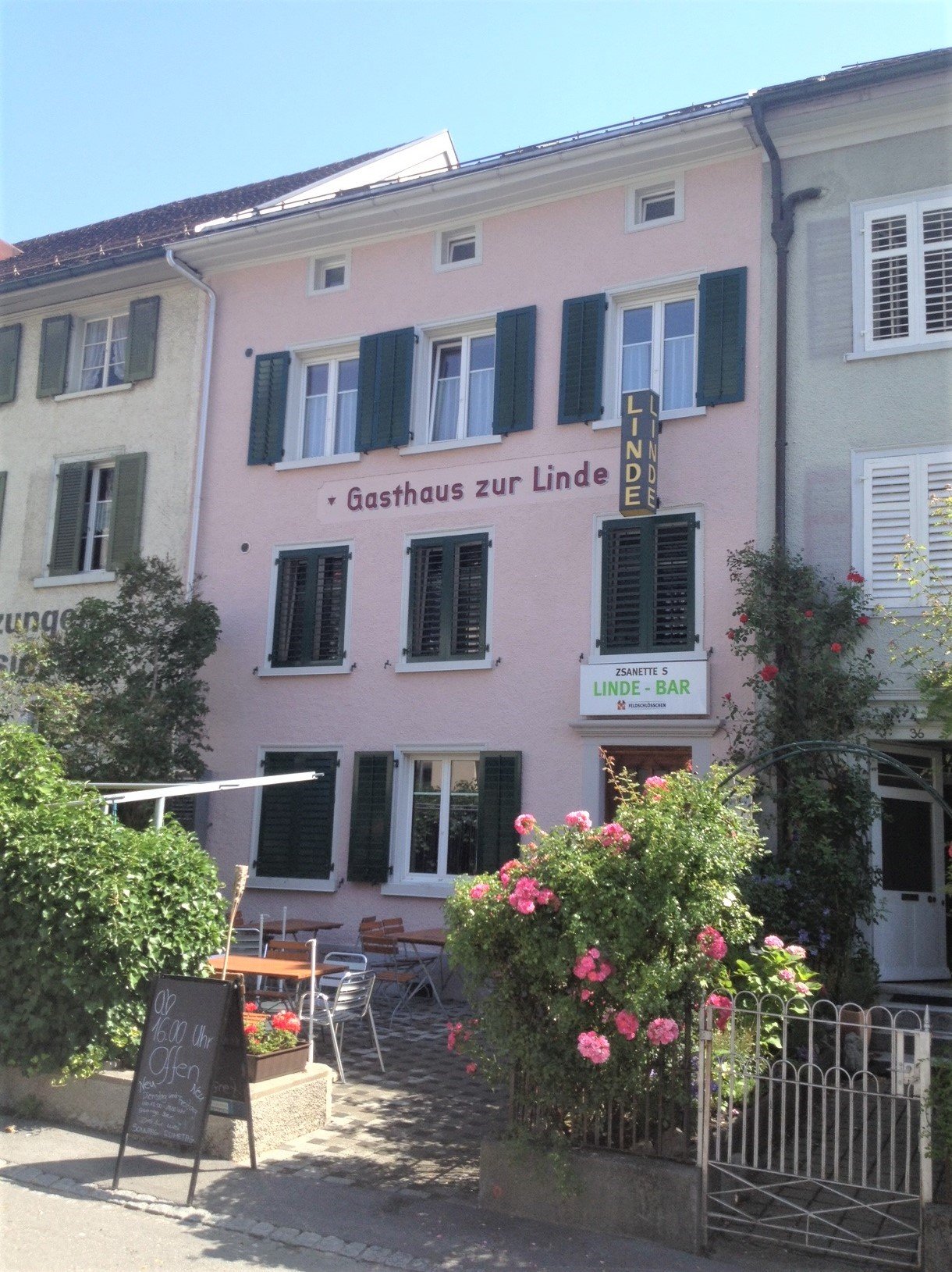 pink building, green shutters, outdoor seating, sign reading Gasthaus zur Linde, flowers, gate