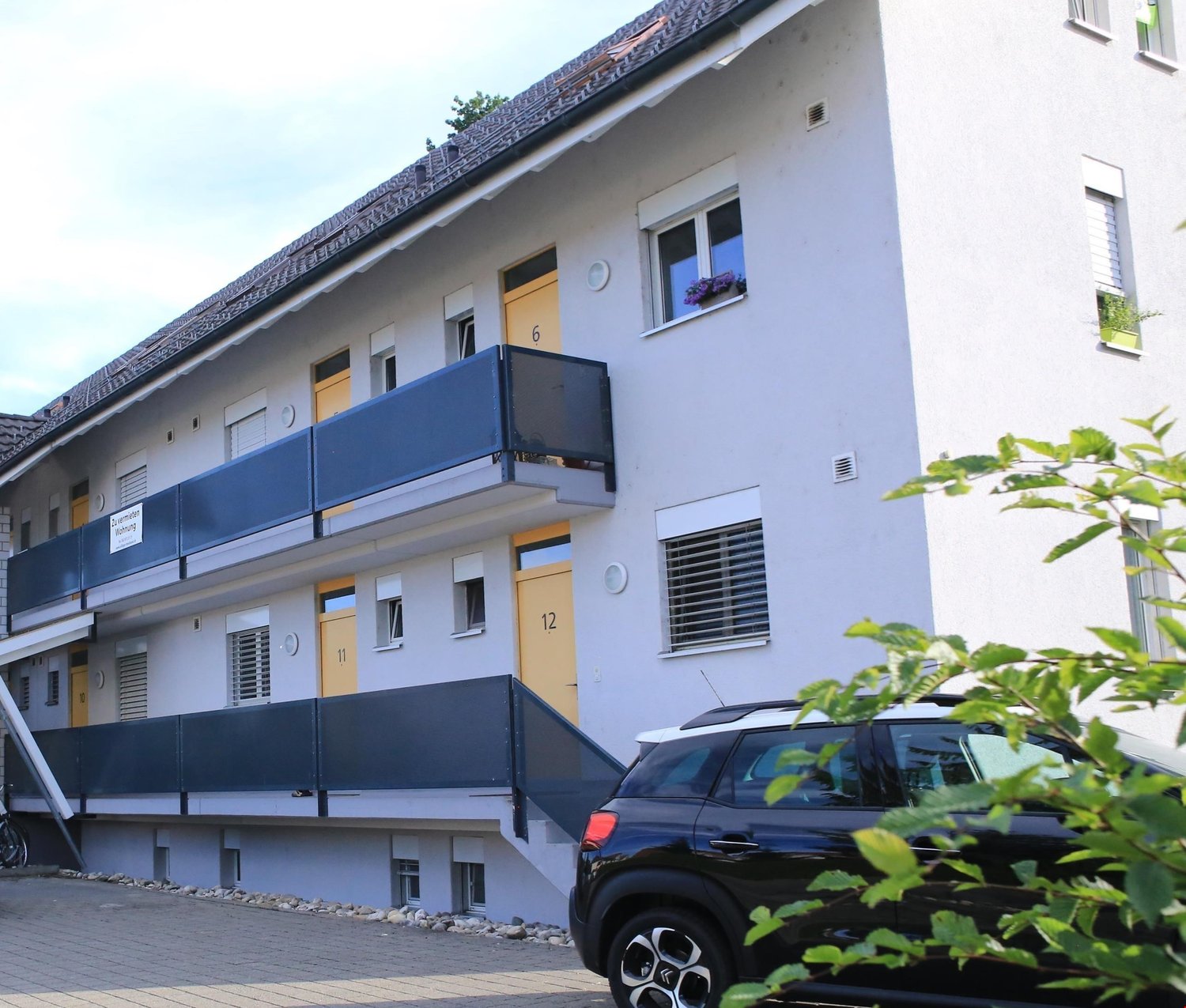 3-story apartment building with white walls, blue balconies, and numbered entrances. A black car is parked in front of the building.