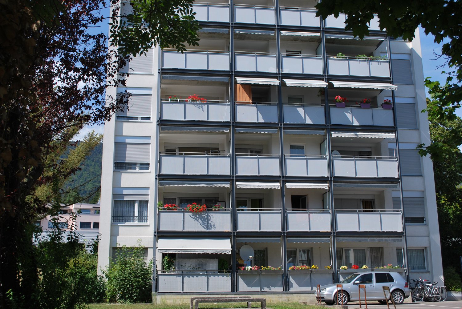 apartment building, multiple balconies, cars parked outside, greenery around the building