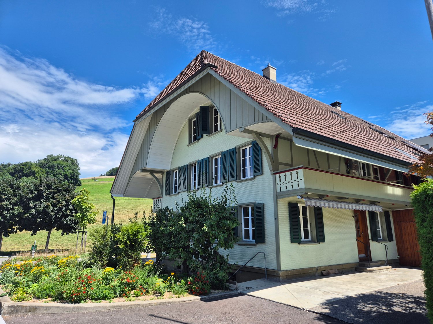 2 story house, brown roof, white walls, several windows, porch, garden with flowers, rural background