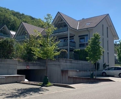 three story house, brown roof, gray concrete barrier, car in front, trees, mountains in background