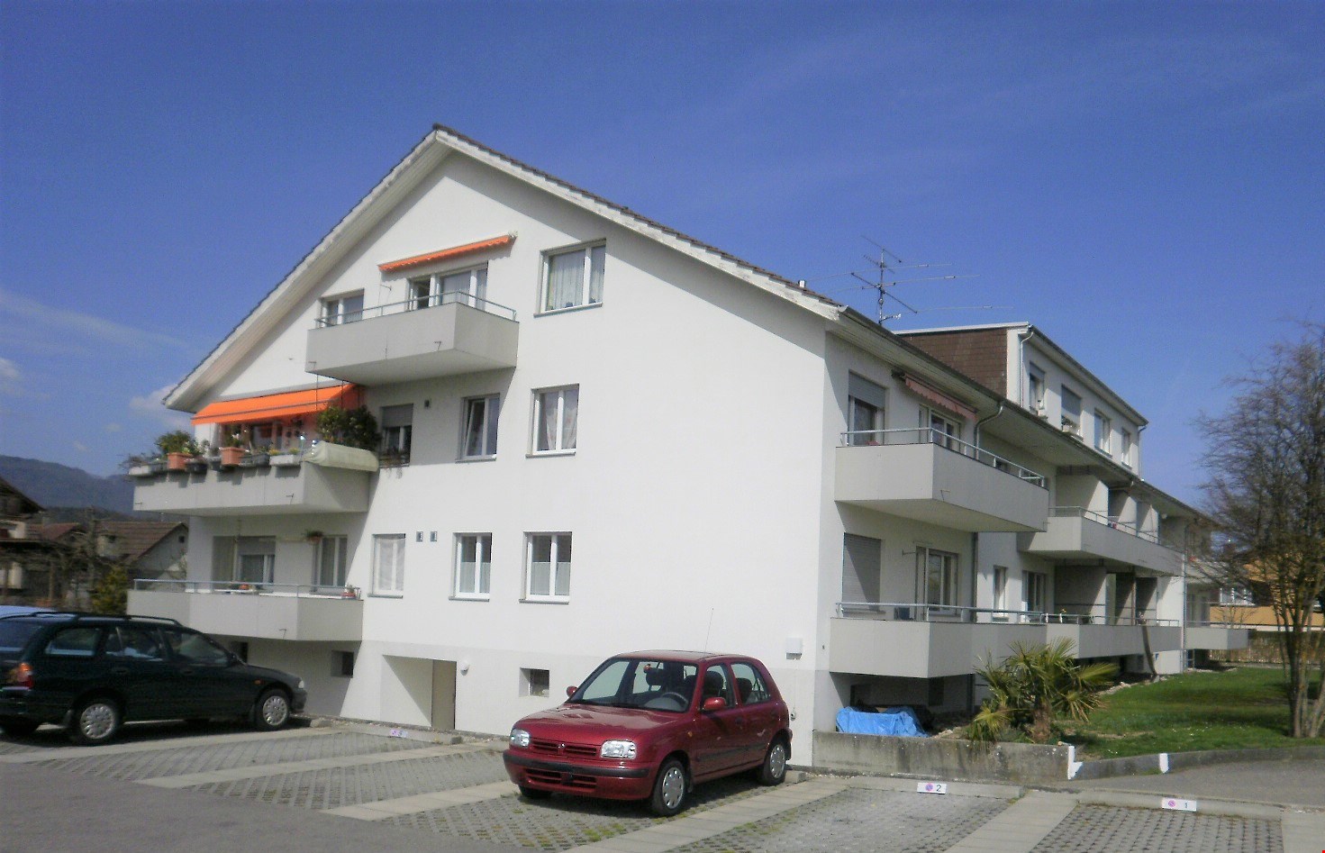 Multi-story apartment building, multiple balconies, white exterior, two cars parked in front.