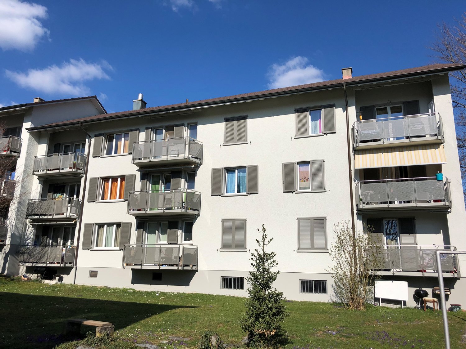 White apartment building with multiple balconies, brown roof, multiple windows, green grass in front
