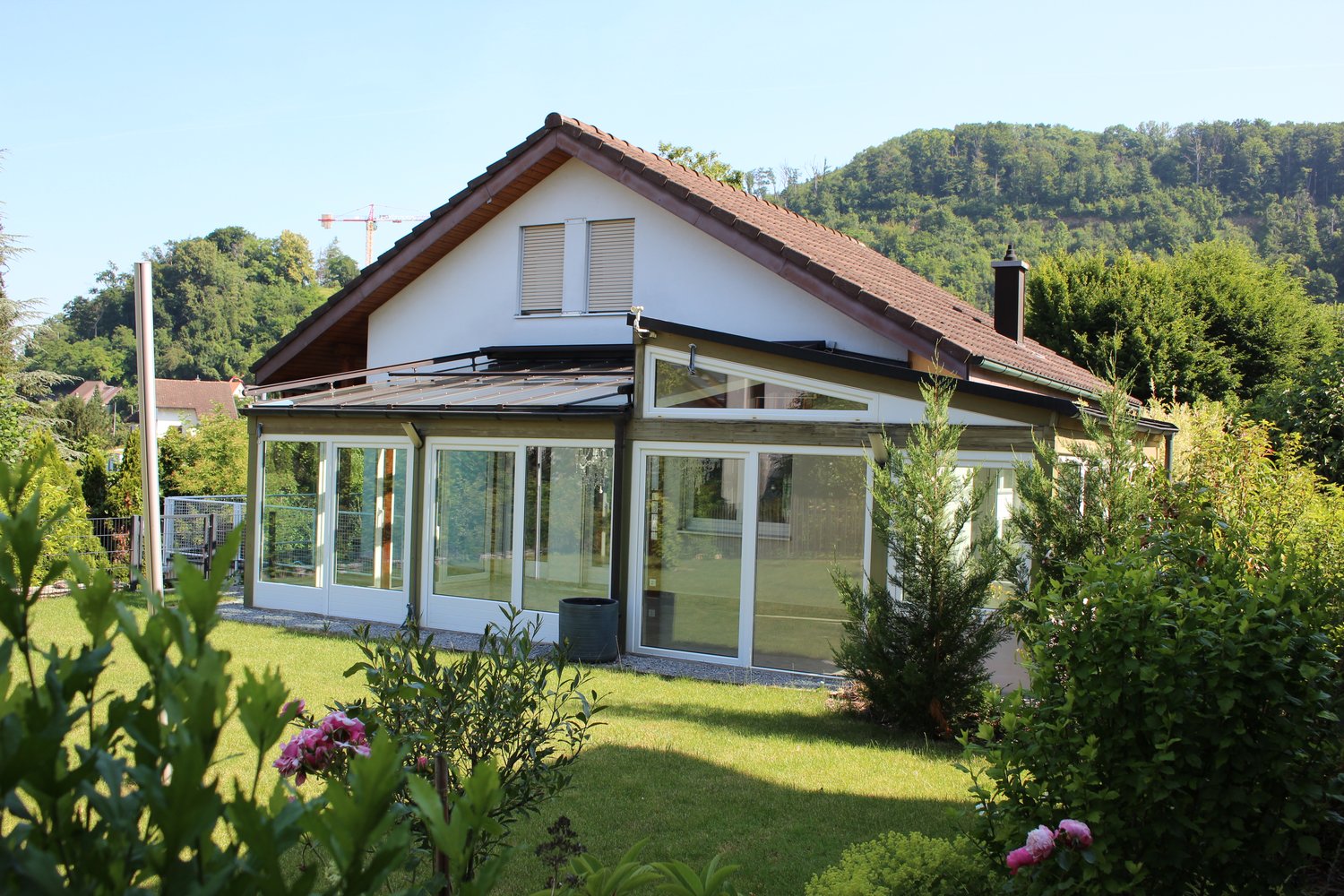 Two-story house with a tiled roof, white exterior walls, and large glass windows. The house has a covered patio or balcony area with glass walls. The house is surrounded by a well-maintained garden with lush greenery and flowering plants.