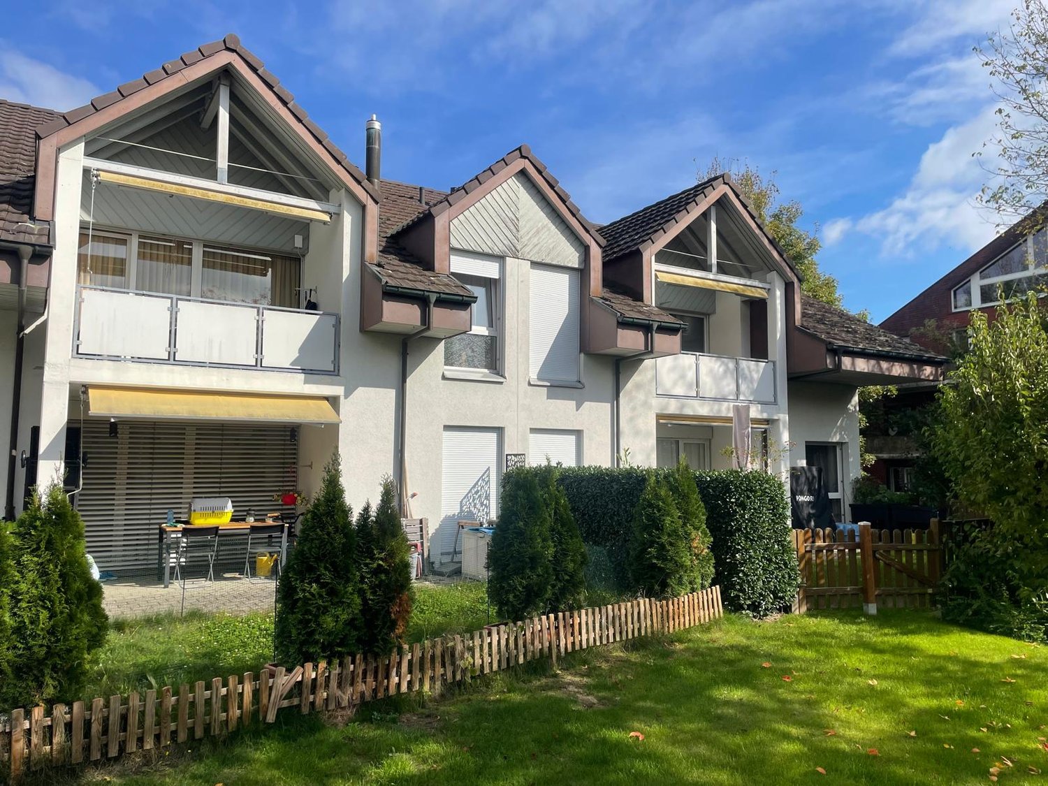 white painted building, multiple balconies, brown roof, fenced garden, wooden fences, trees
