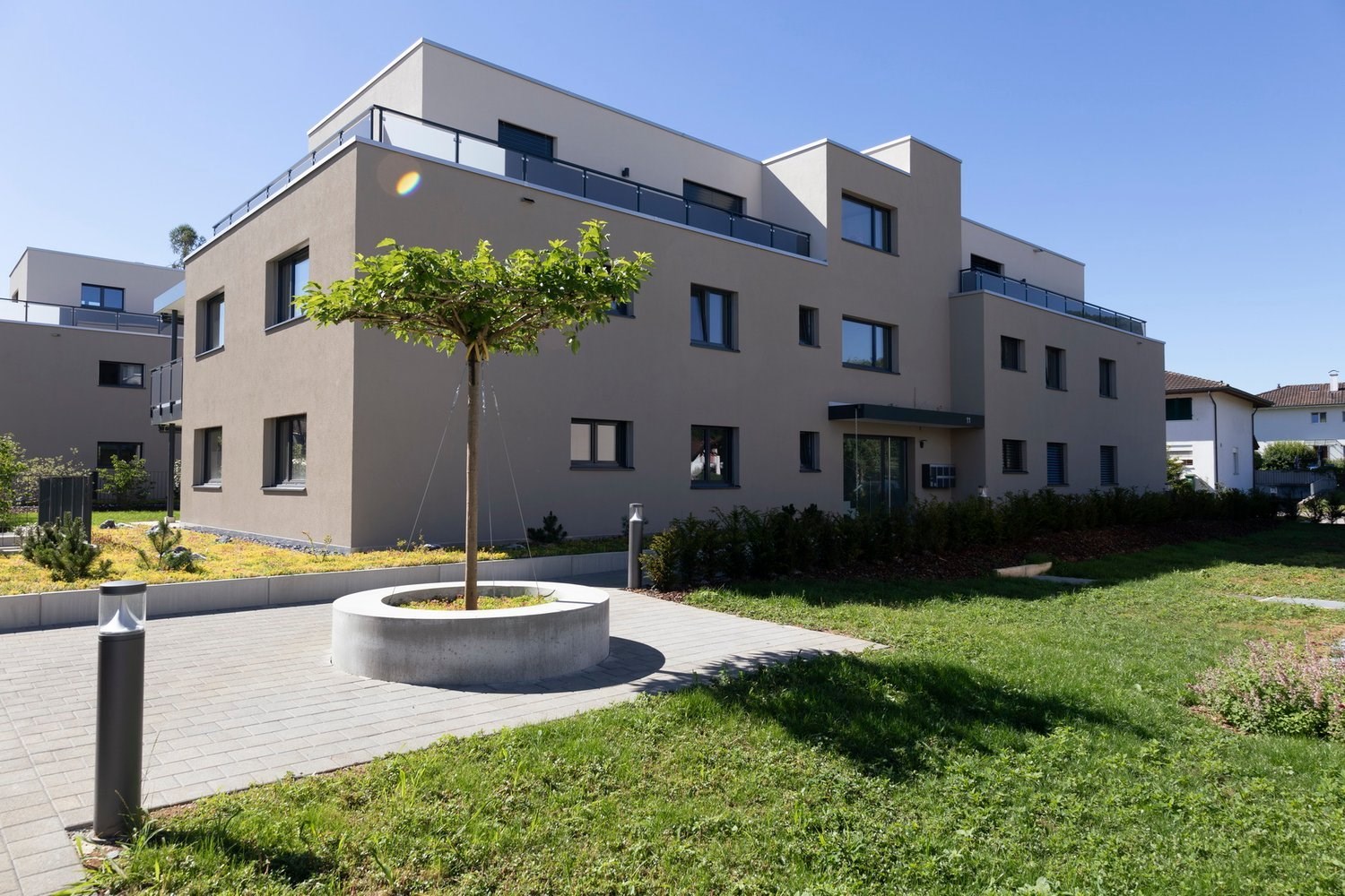 Modern three-story apartment building, beige facade, large windows, central tree, paved walkway, front yard with lawn and shrubs