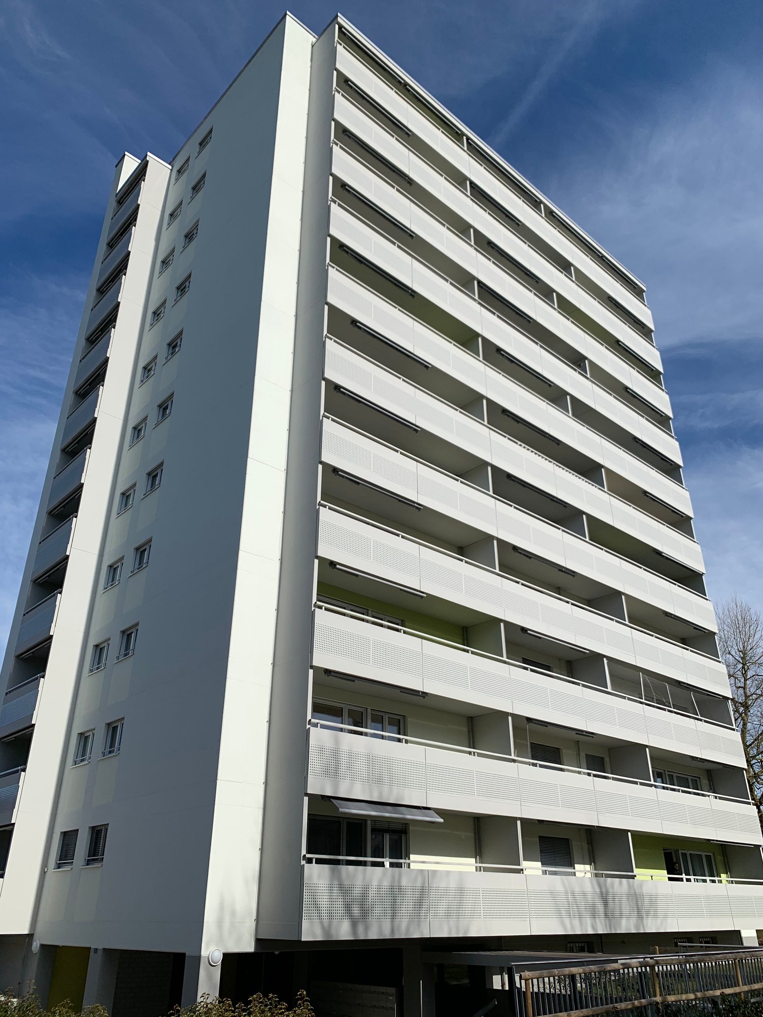 Multi-story apartment building with white exterior, balconies on each floor, and a blue sky in the background.