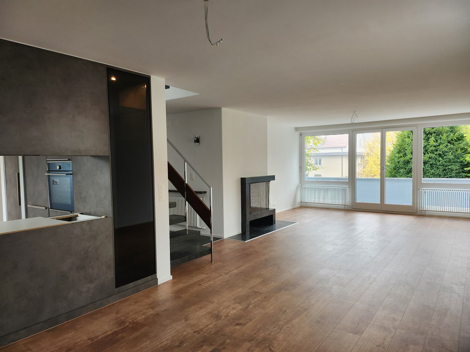 Open-plan living room with modern decor and wooden flooring, fireplace, kitchen island with oven and sink, large glass doors leading to a balcony, white walls, ceiling lamps, staircase with handrail