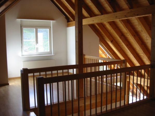 The image shows an attic or loft space with exposed wooden beams and rafters in the ceiling. There is a window providing natural light, and a wooden railing or balcony overlooking the space below.