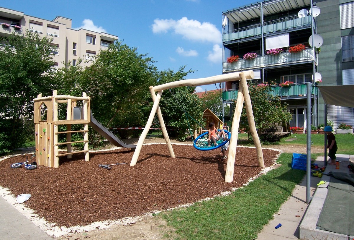 The image shows a residential area with apartment buildings and a playground. The playground has a wooden structure with a slide and swings. The buildings have balconies and there are trees and greenery surrounding the area.