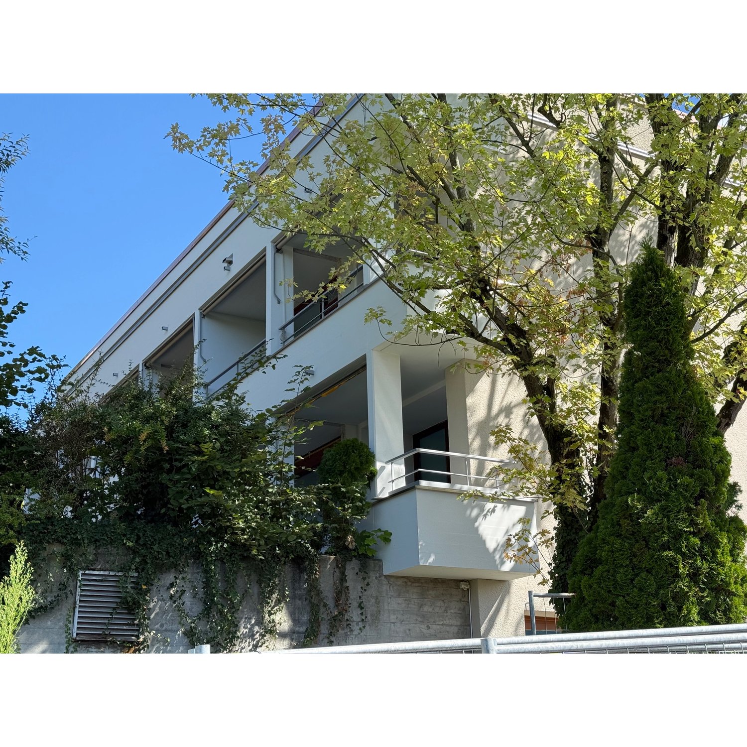 white building, multiple floors, balcony, trees in front, window air vents on side wall
