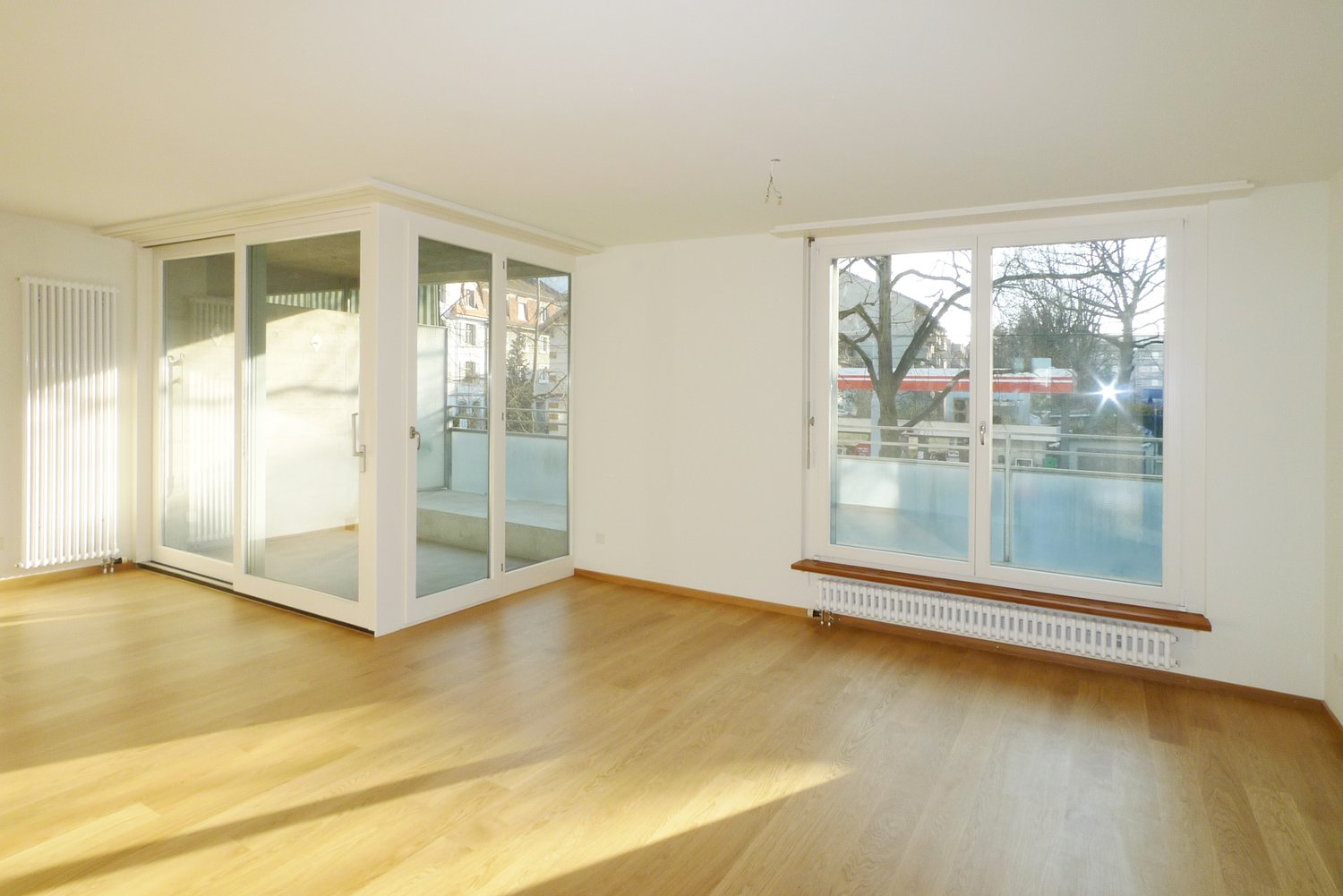 An empty room with glass doors leading to the balcony, wooden floor, white walls and large windows