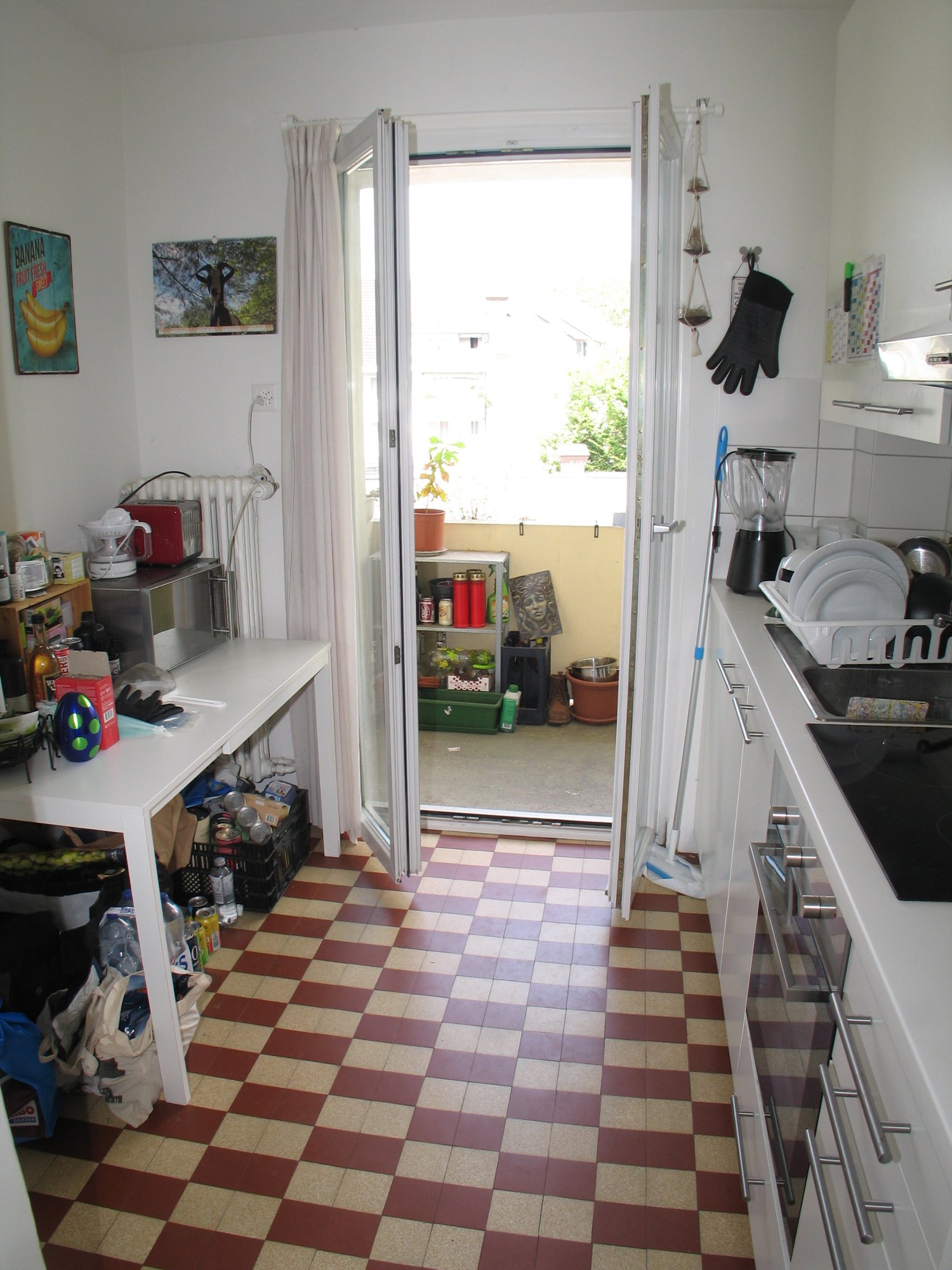 Kitchen with red and white checkered floor, open sliding door to balcony, shelves with canned goods and potted plants, small work table, microwave, toaster, counter with food processor and bottles, oven, stovetop, plate rack, broom, stack of plates