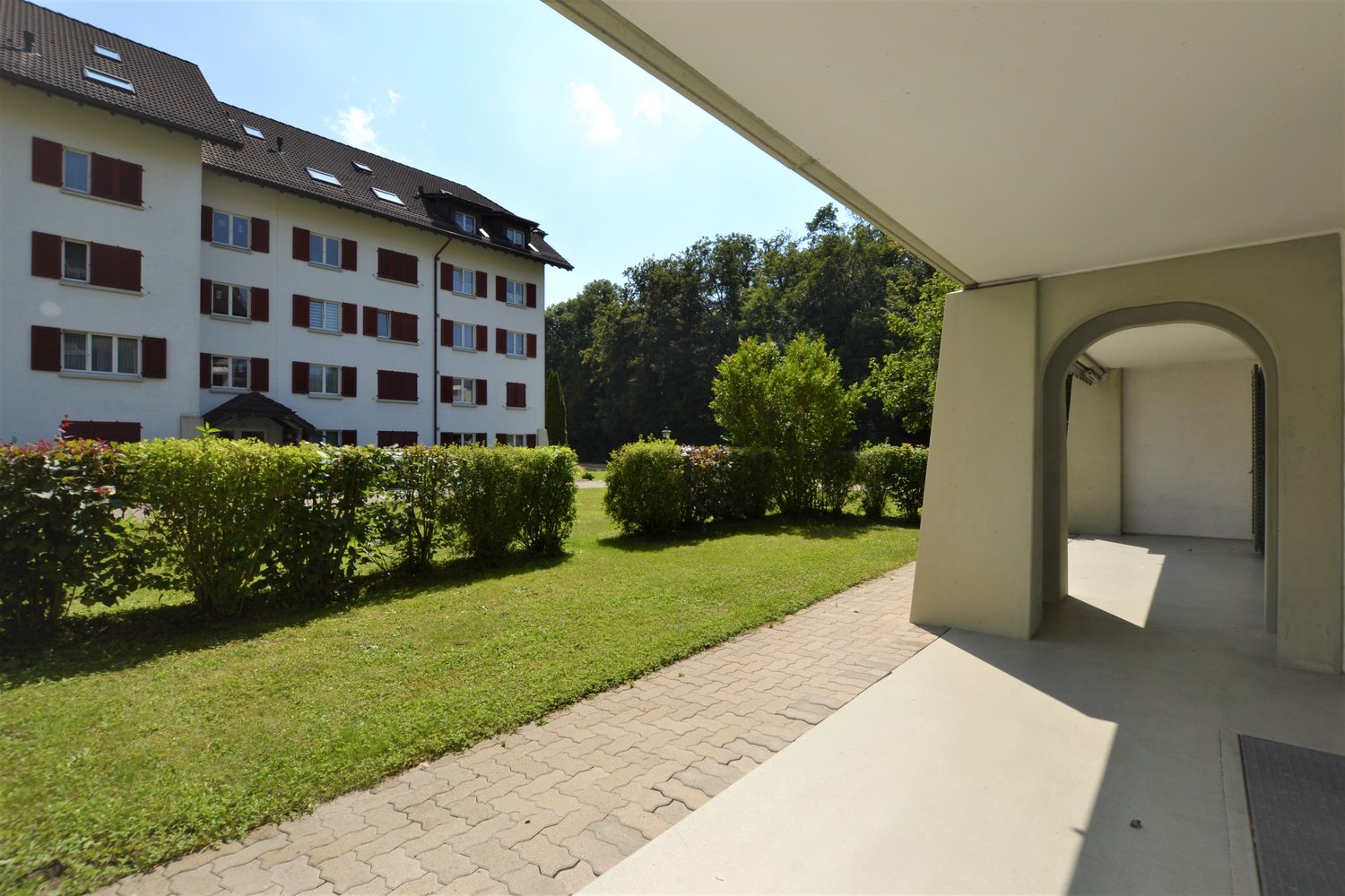 Apartment building with 4 floors, white exterior, red shutters, brick pathway, bushes, grass lawn, trees in the background.