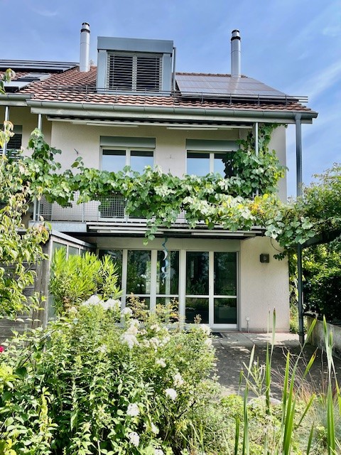 2 story house, white walls, red roof, large glass windows, covered balcony, plants and vines over balcony, solar panels on roof
