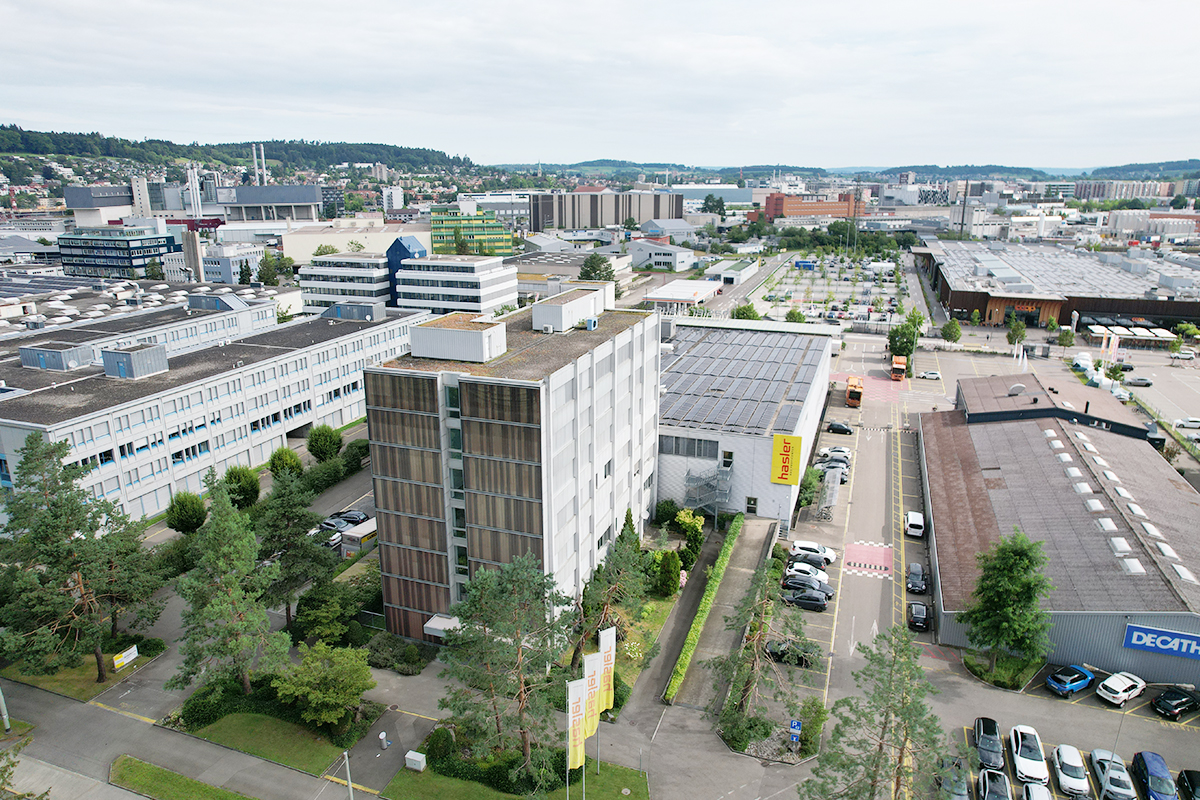 Aerial view of an urban area with buildings, parking lots, trees, and distant hills.
