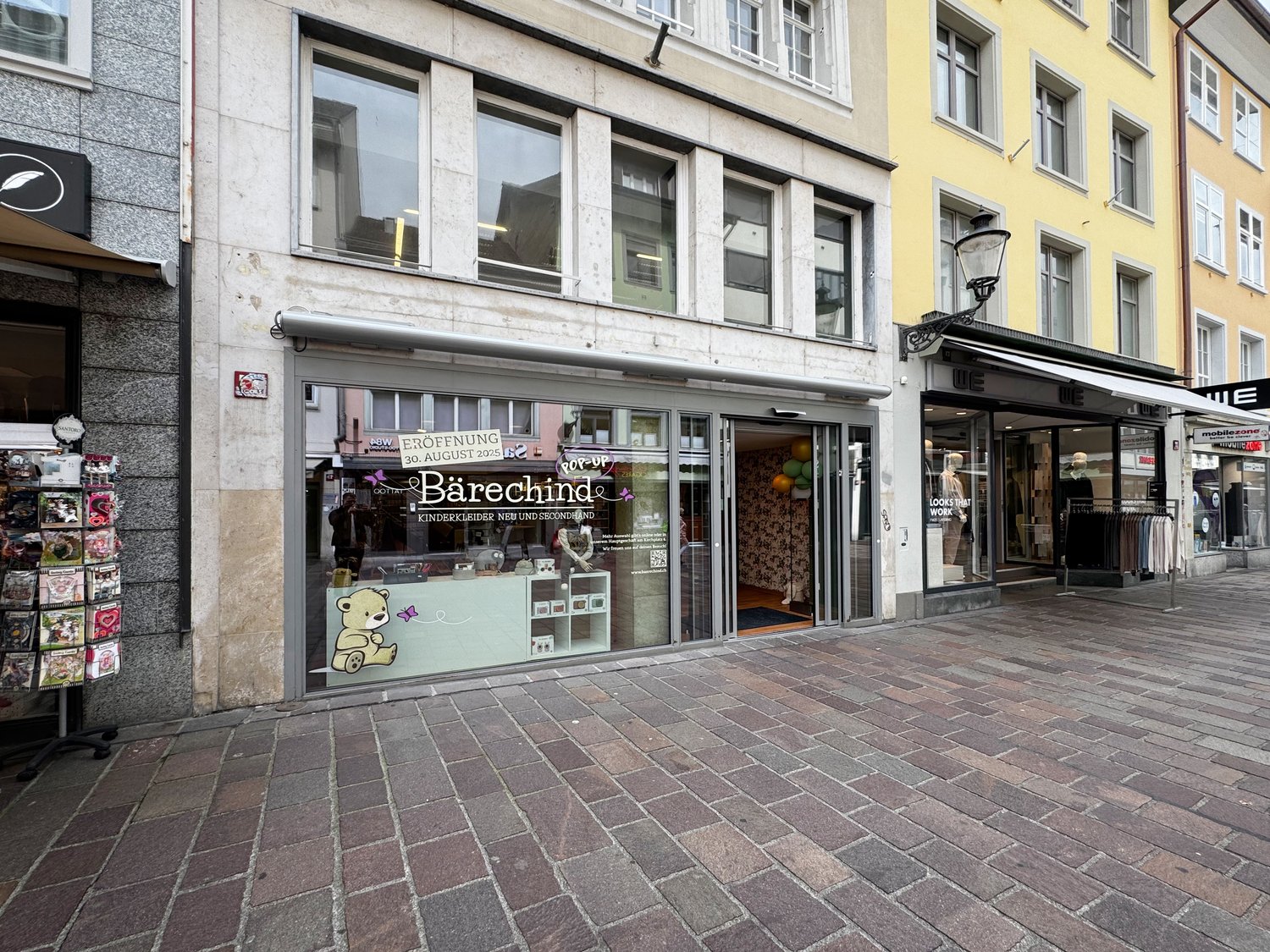 The storefront is a multi-story building with large windows, glass doors, and awnings. Inside the storefront, there is a shop with a display of products. The storefront is located in a busy street with other shops and a brick sidewalk.